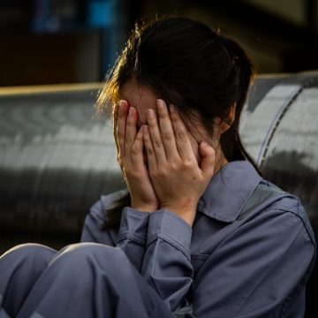 Distressed sad Asian factory worker woman in blue uniform sitting on floor crying covering face with hands feeling hopeless depressed exhausted tired alone LEGAL REGULATORS MUST HOLD LAW FIRMS ACCOUNTABLE FOR HARASSMENT